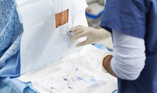 Regional-Anesthesia 1-min Gloved hand inserting epidural anesthesia syringe at patient's spine; clinician standing in front of tray medical supplies