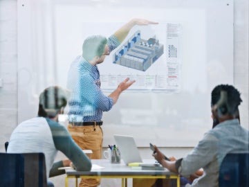 Man in blue checkered shirt and khaki pants explains a supply room schematic to two men seated at a table
