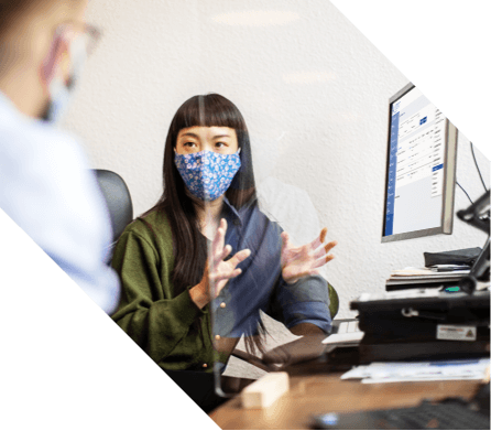 A woman at a desk with a mask on talking to a man