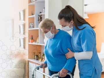 Female caregiver in blue scrubs helping a gray-haired woman in a blue sweater use a walker